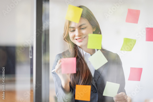 Business Asian female employee with many conflicting priorities arranging sticky notes commenting and brainstorming on work priorities colleague in a modern office.