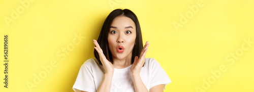 Close up of surprised asian girl say wow, stare at camera amzed with hands near face, standing over yellow background photo