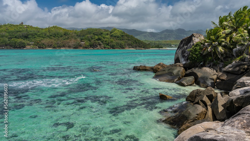 Corals are visible through the clear turquoise ocean water. On the shore there are picturesque granite boulders, lush palm trees. Green hills against the sky and clouds. Seychelles.