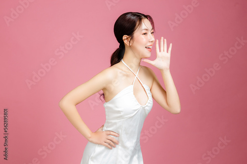 Young Asian woman gathered in ponytail with natural makeup on face have plump lips and clean fresh skin wearing white camisole open mouths raising hands announcement on isolated pink background. photo