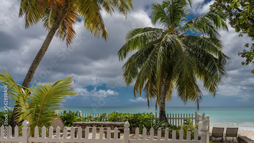 The relaxation area on the tropical beach is surrounded by a white fence. A wooden table, benches, and deck chairs are visible. Palm trees against a blue sky, clouds, turquoise ocean. Seychelles