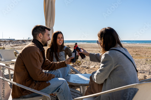 Young friends in warm clothes sitting at table and clinking bottles of drinks while resting during weekend on beach