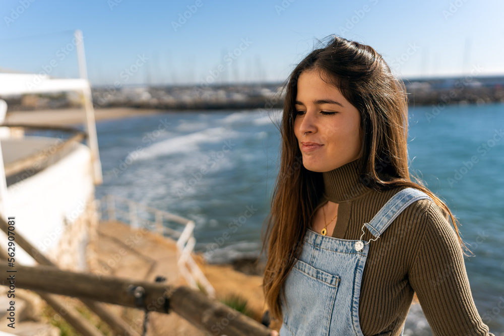 Hispanic female with long hair closing eyes delightfully while standing on pier near rippling sea and enjoying sunshine