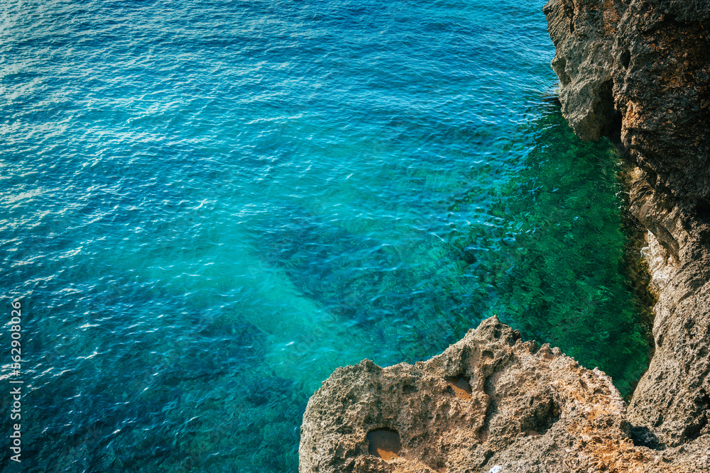 Top view of turquoise, blue water and gray rocks to the right. View from a cliff over the clear sea with plenty of copy space