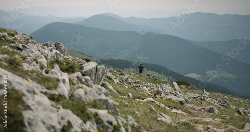 Hiker walking climbin a mountain with hiking poles, brhnd him is a valley and nearby mountains partially covered in thin clouds. photo