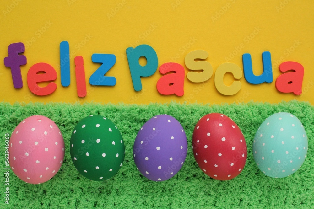 Feliz Pascua, Happy Easter letters on spanish on yellow background and color eggs on green grass. Holiday banner, flatlay creative composition. Greeting card, poster, banner. Top view festive concept