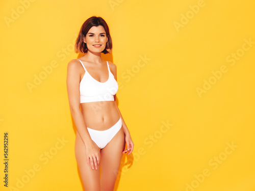 Fashion portrait of young beautiful woman. Attractive carefree blond model wearing pure white lingerie. Hot tanned smiling blonde posing near yellow wall in studio, showing sexy natural body