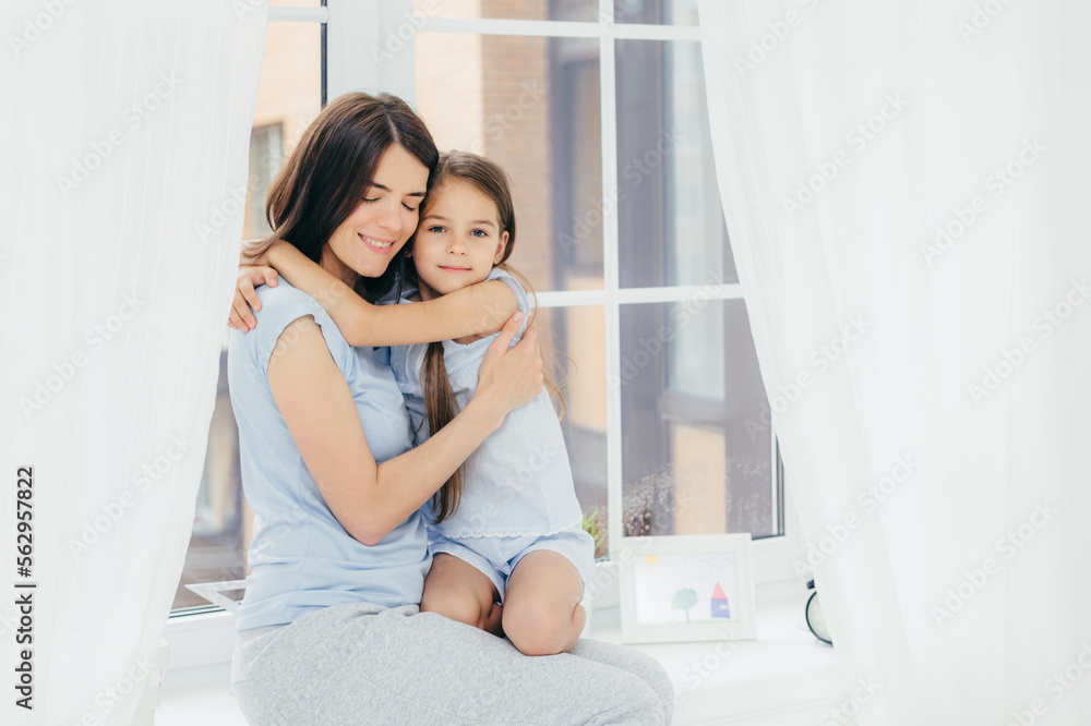 Lovely small child with pleasant appearance embraces her mother, expresses love and good feeling or attitude, sit on window sill, enjoy domestic atmosphere and togetherness. Parenthood concept Lovely small child with pleasant appearance embraces her mother, expresses love and good feeling or attitude, sit on window sill, enjoy domestic atmosphere and togetherness. Parenthood concept