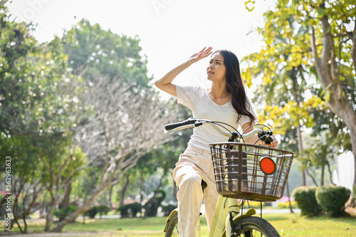 Tired young Asian woman is on her bike, taking a rest, looking at the nature view of the public park.