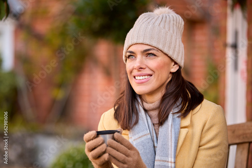Woman Wearing Hat And Scarf With Hot Drink Sitting On Bench Outside House In Autumn 