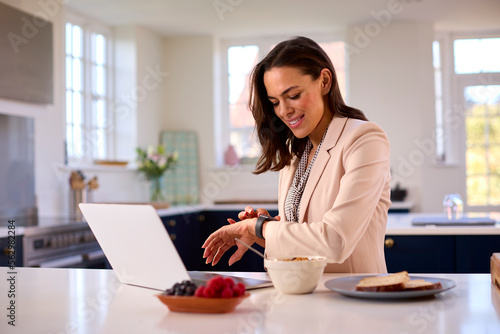 Businesswoman At Home With Laptop On Kitchen Counter Eating Breakfast Checking Smart Watch