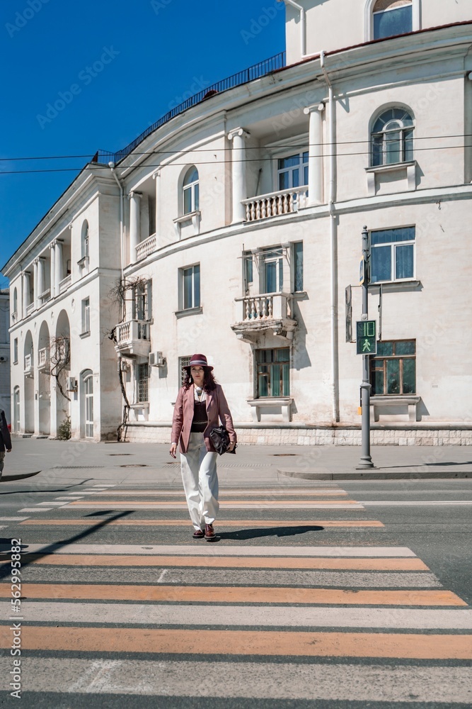 Woman city road crossing. Stylish woman in a hat crosses the road at a pedestrian crossing in the city. Dressed in white trousers and a jacket with a bag in her hands.