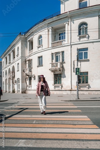 Woman city road crossing. Stylish woman in a hat crosses the road at a pedestrian crossing in the city. Dressed in white trousers and a jacket with a bag in her hands. © svetograph
