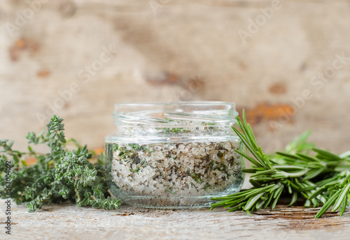Homemade sea salt scrub with olive oil, rosemary and thyme (bath salt, foot soak, facial or hair mask) in a glass jar. Natural beauty treatment and spa recipe. Copy space, old wooden background.