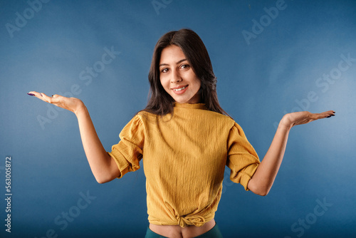 Portrait of cute dark-haired woman wearing casual top isolated over blue background shrugging and throwing hands aside with clueless. Not sure what to do. Looking at the camera.