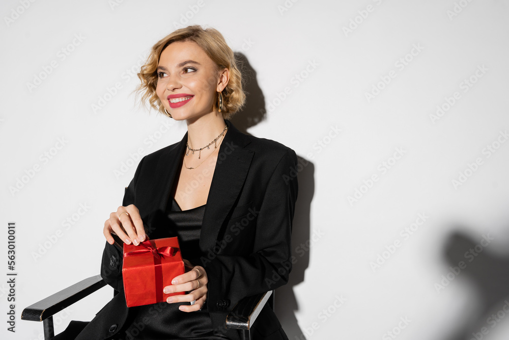 positive woman in black blazer and slip dress holding wrapped present while sitting on grey. positive woman in black blazer and slip dress holding wrapped present while sitting on grey.