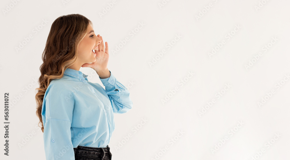 Hey! Cheerful young lady in blue shirt is shouting and holding hand near her open mouth on light white background Hey! Cheerful young lady in blue shirt is shouting and holding hand near her open mouth on light white background