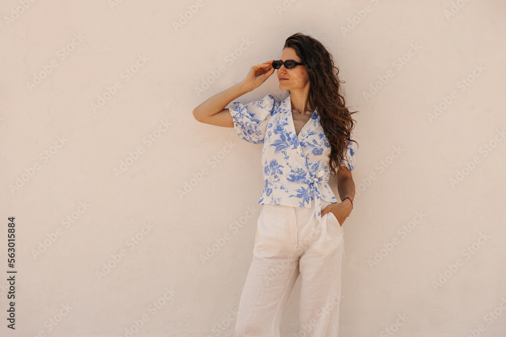Fashionable young caucasian woman in sunglasses posing with hand in pocket near light wall. Brunette hair model wears white shirt and casual clothes. Concept summer vacation. Fashionable young caucasian woman in sunglasses posing with hand in pocket near light wall. Brunette hair model wears white shirt and casual clothes. Concept summer vacation.