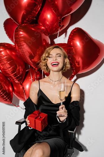 happy woman in black slip dress holding glass of champagne and present while sitting near red balloons on grey.