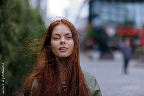 Portrait of a beautiful young girl in the city looking into the camera with red flying hair in a green raincoat in the city against a background of bamboo in spring, lifestyle in the city