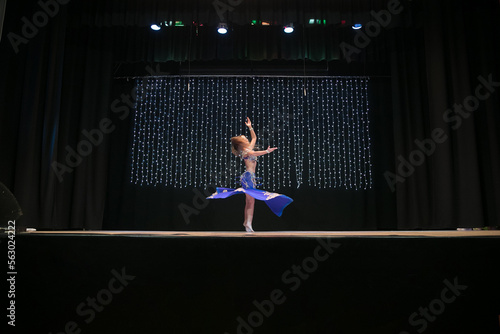 Bild auf Leinwand A young dancer with long hair in an oriental costume with a multi-colored shawl poses and dances a belly dance on a concert stage