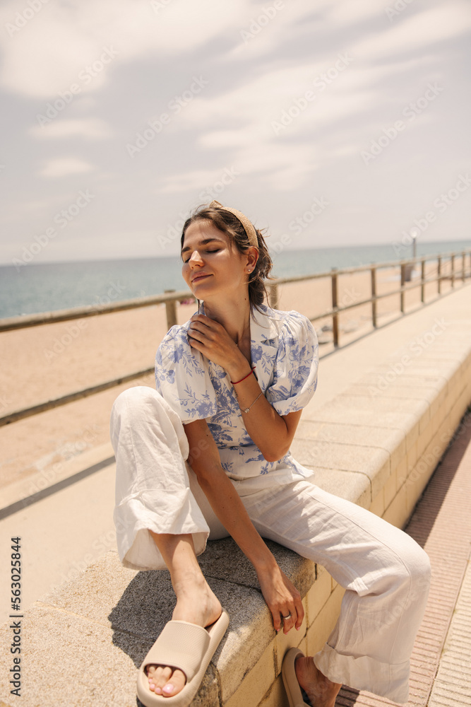 Tender young caucasian woman is sitting on embankment by sea, posing with eyes closed in sunny weather. Brunette wears casual clothes. Concept of relaxation, rest. Tender young caucasian woman is sitting on embankment by sea, posing with eyes closed in sunny weather. Brunette wears casual clothes. Concept of relaxation, rest.