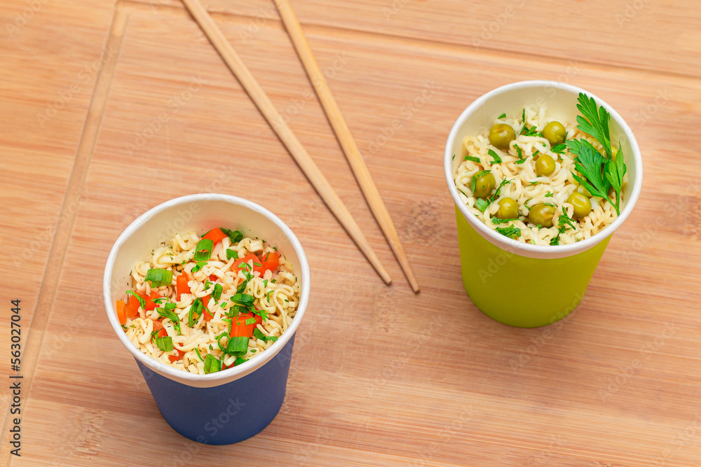 Prepared Instant Noodles with Green Pea, Carrot and Greens in Blue and Green Cups with Chopsticks on Wooden Cutting Board. Quick Lunch or Unhealthy Fast Food Prepared Instant Noodles with Green Pea, Carrot and Greens in Blue and Green Cups with Chopsticks on Wooden Cutting Board. Quick Lunch or Unhealthy Fast Food