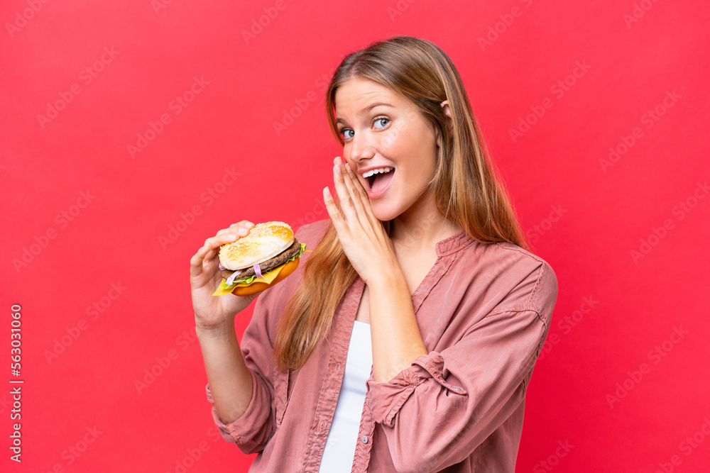 Young caucasian woman holding a burger  isolated on red background whispering something