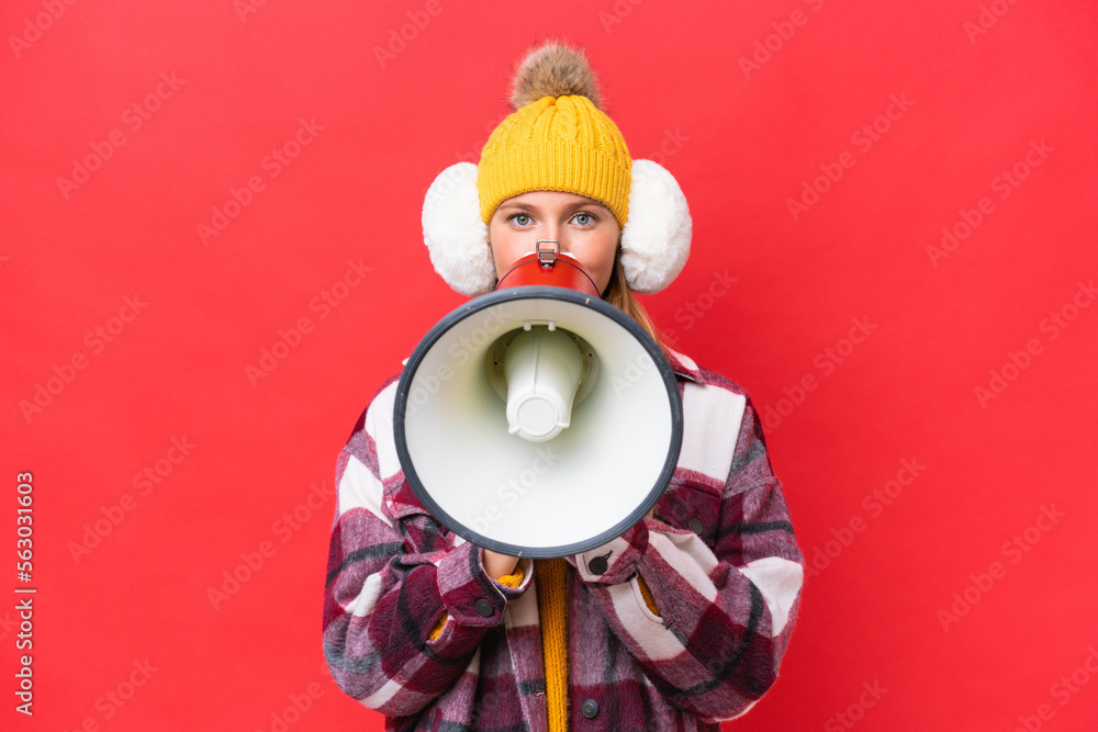 Young beautiful woman wearing winter muffs isolated on red background shouting through a megaphone