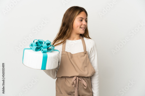Little girl holding a big cake over isolated white background looking to the side and smiling