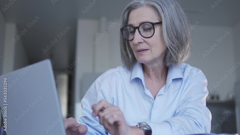 Mature woman political scientist conducting researches, looking for information in internet, working on laptop sitting at home office, making notes at notepad Mature woman political scientist conducting researches, looking for information in internet, working on laptop sitting at home office, making notes at notepad