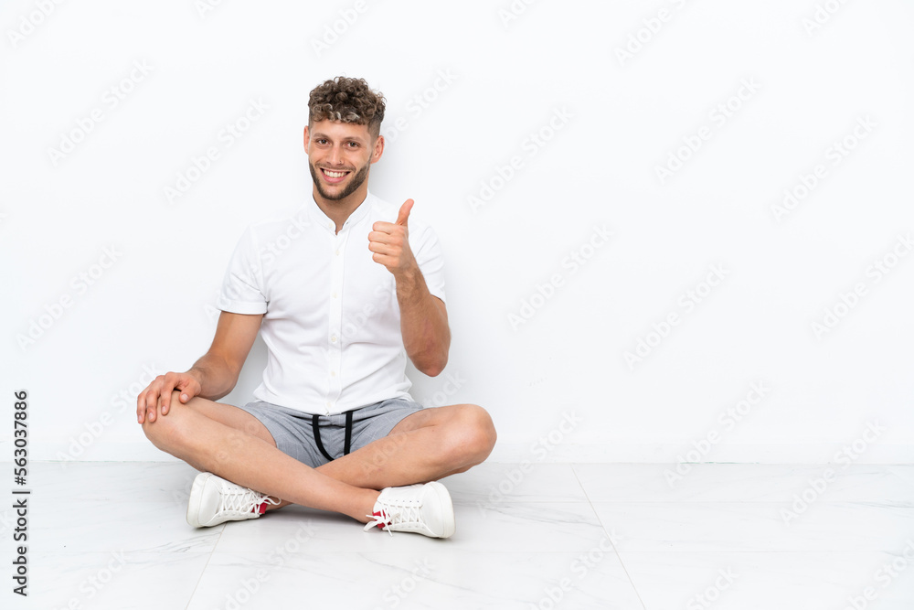 Young blonde man sitting on the floor isolated on white background with thumbs up because something good has happened Young blonde man sitting on the floor isolated on white background with thumbs up because something good has happened