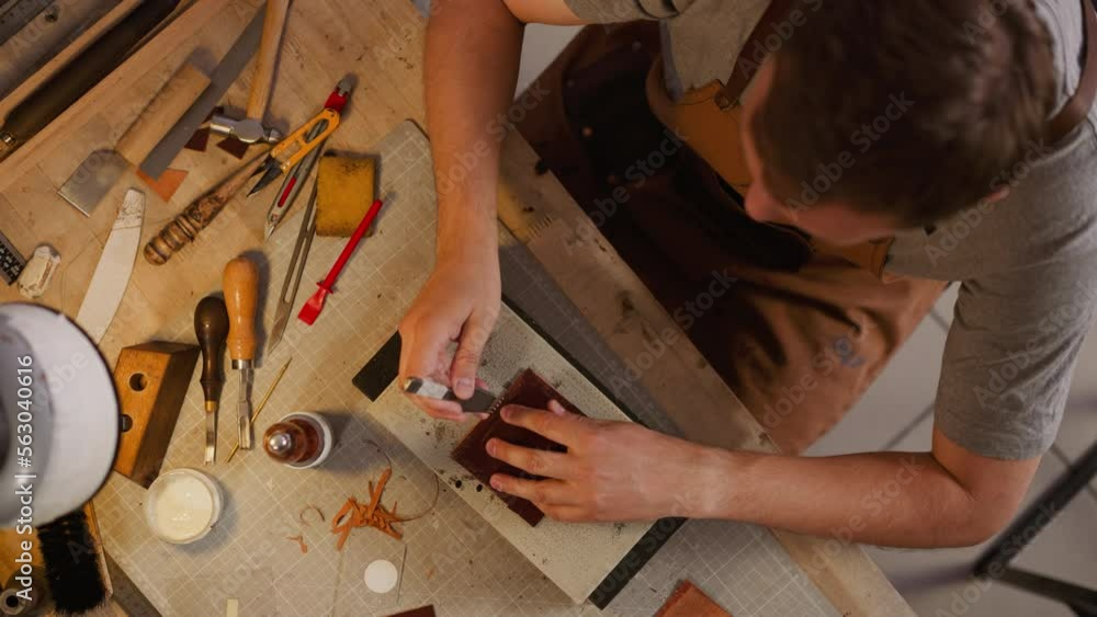 Worker pierces leather piece with awl and special round hammer at table in studio view from above. Tradition of handicraft leather items creation Worker pierces leather piece with awl and special round hammer at table in studio view from above. Tradition of handicraft leather items creation