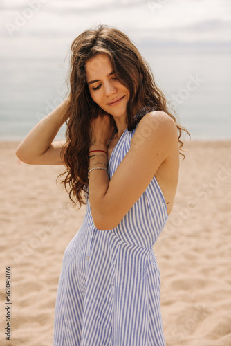 Nice young caucasian girl stands on beach, enjoys time on summer evening. Brunette woman wears striped dress with sea in background. Concept enjoying mood.