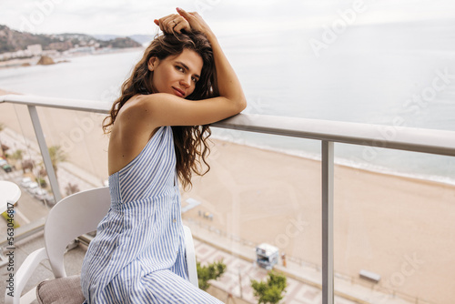 Beautiful young caucasian girl wears striped dress sitting chair on balcony with beach in background. Brown hair woman spends vacation sunny country. Concept summer vacation.