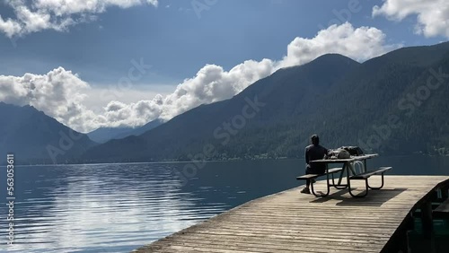 A female hiker sits at a picnic bench on a wooden dock overlooking a beautiful rippling lake with forest covered mountains and clouds in the background - Lake Crescent, Washington, USA photo