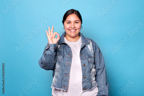 Young curvy latina woman wearing denim jacket and hoop earrings, smiling showing okay sign looking at camera isolated on blue background. Copy space. photo