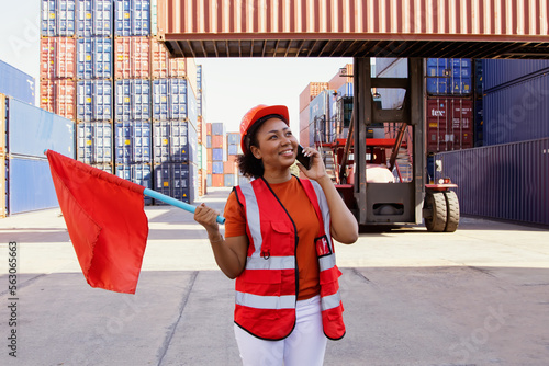 African american female worker holding red flag giving traffic signal passing through container loading yard safe transportation caution using smartphone talking worker working together as a team.