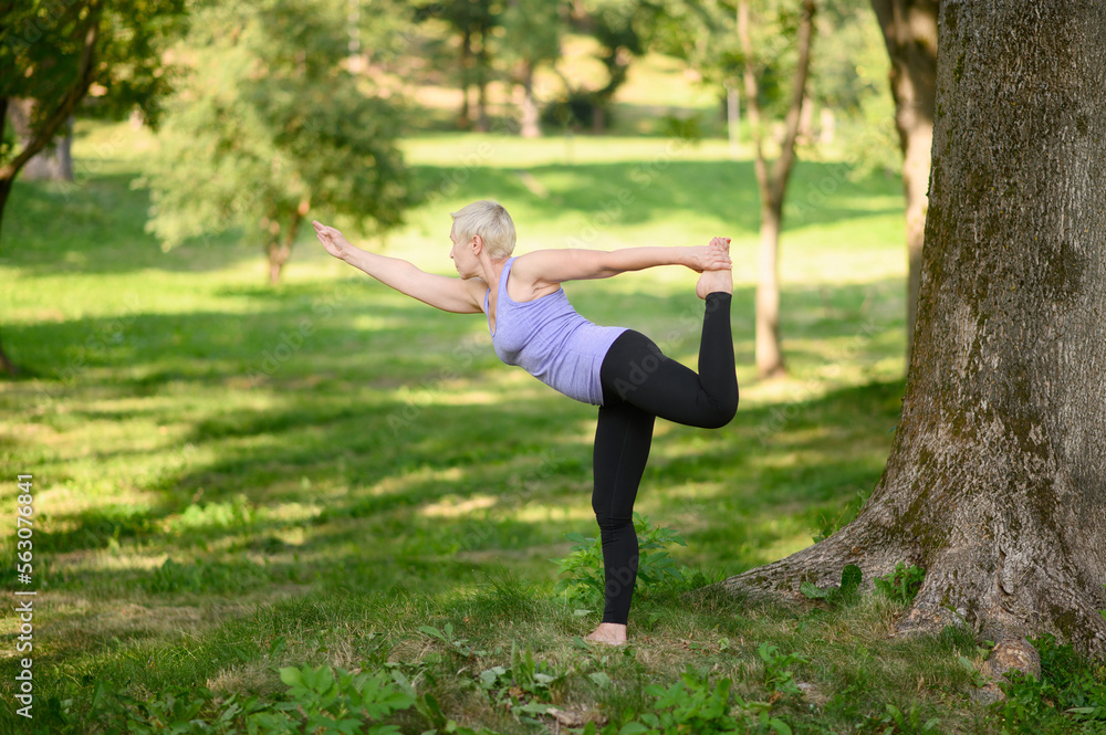 A middle-aged woman practices yoga doing the Three Warrior Postures exercises holding her bent leg with one hand in the park.The concept of an active lifestyle, body care, training, stretching, yoga. A middle-aged woman practices yoga doing the Three Warrior Postures exercises holding her bent leg with one hand in the park.The concept of an active lifestyle, body care, training, stretching, yoga.