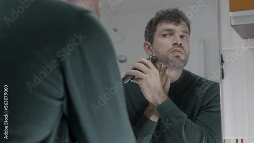 Young attractive man shaving in front of mirror in bathroom electric shaver getting ready grooming taking care of aspect hygiene concept care routine