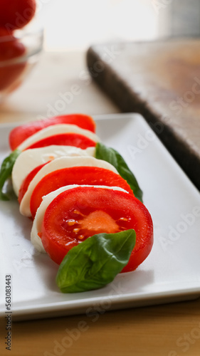 Vertical close up view of a fresh caprese salad over a white rectangular plate  kitchen knife and a bowl of fresh tomatoes over a wooden table