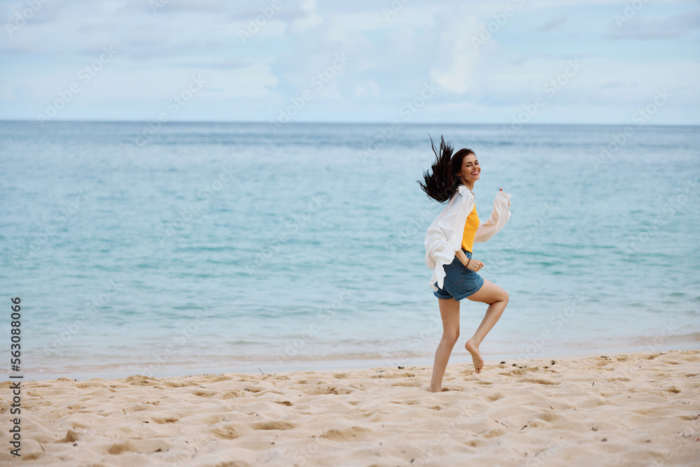 Fototapeta premium Sports woman runs along the beach in summer clothes on the sand in a yellow T-shirt and denim shorts white shirt flying hair ocean view, beach vacation