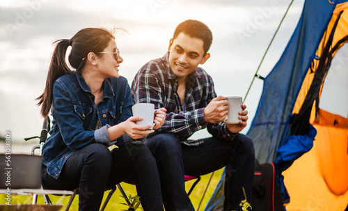 Happy Asian young couple sitting on picnic chair drinking tea and coffee while tent camp lakeside at parks outdoors on vacation holiday. Adventure lifestyle of man and woman with camping in nature.