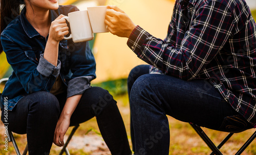 Happy Asian young couple sitting on picnic chair drinking tea and coffee while tent camp lakeside at parks outdoors on vacation holiday. Adventure lifestyle of man and woman with camping in nature.