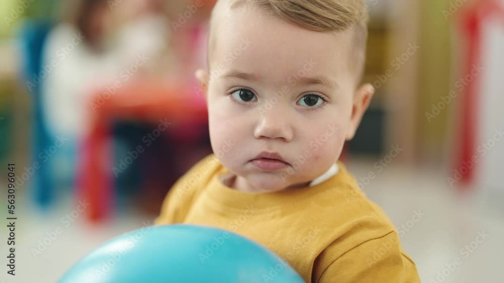 Adorable blond toddler holding ball standing with relaxed expression at kindergarten Adorable blond toddler holding ball standing with relaxed expression at kindergarten
