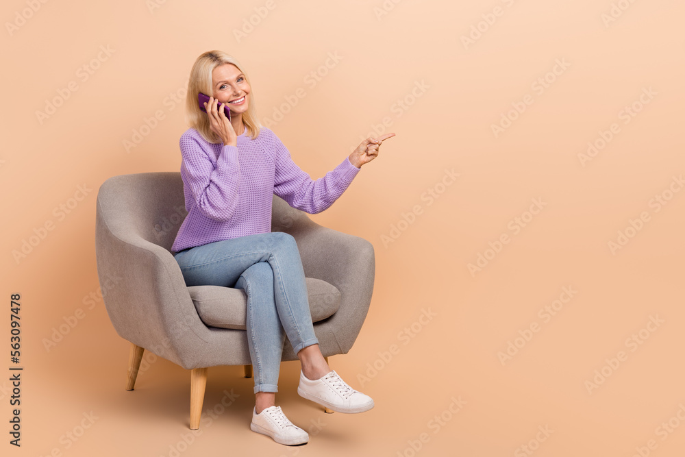 Full size photo of nice woman dressed purple sweater jeans on armchair talking directing empty space isolated on beige color background Full size photo of nice woman dressed purple sweater jeans on armchair talking directing empty space isolated on beige color background