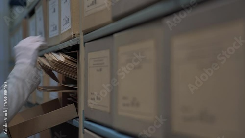 Worker in cotton gloves and grey jacket looks for archive criminal case record in archive cardboard box on shelf in storehouse closeup
