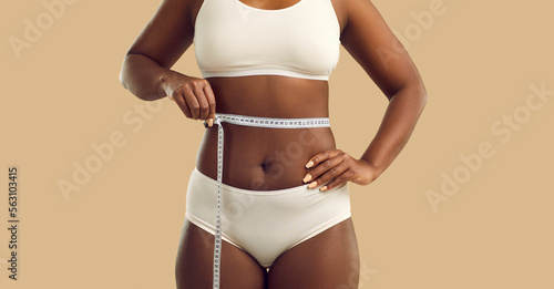 Slika na platnu Plus size woman holding measuring tape on waist on beige color background, cropped shot