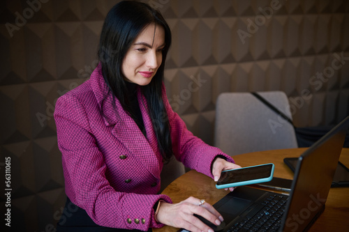 Successful confident 40-45 years old middle-aged Caucasian woman  entrepreneur in casual wear  holding mobile phone and typing text on laptop in a cozy office interior. People  online business concept