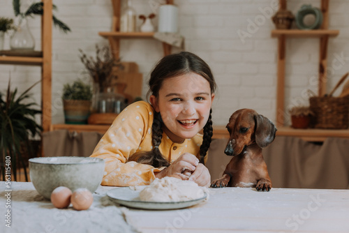 cute little girl at home in the kitchen bakes cookies with her pet dog dachshund. child with a pet in the kitchen